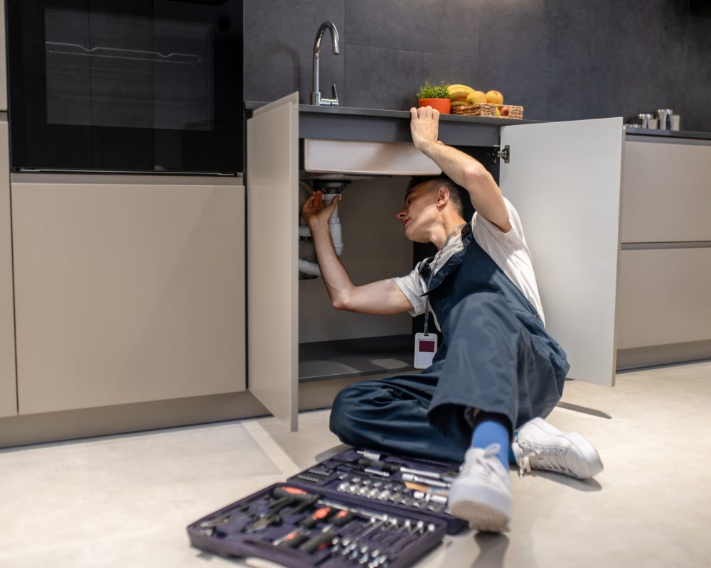 Household repair. Middle aged man in blue jumpsuit reclining professionally inspecting pipe touching hand under sink in stylish modern kitchen
